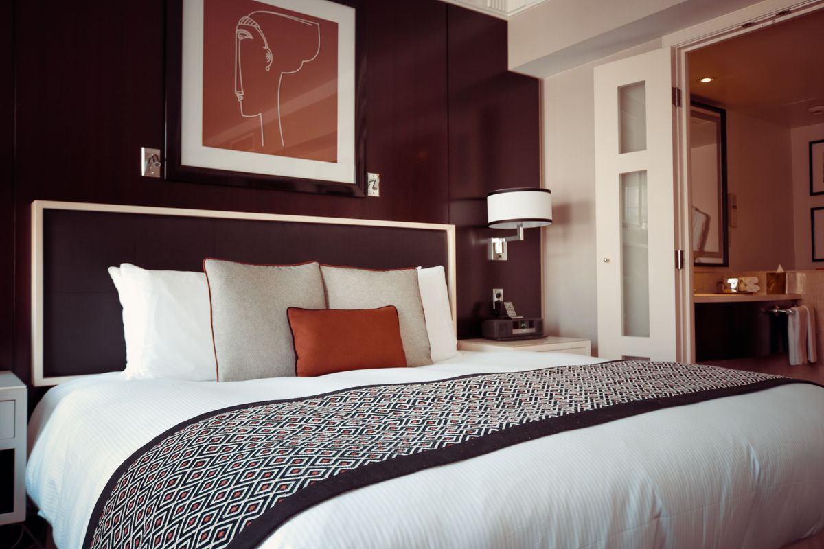 A made bed in a warm, dark walled boutique hotel room with framed line art above the headboard